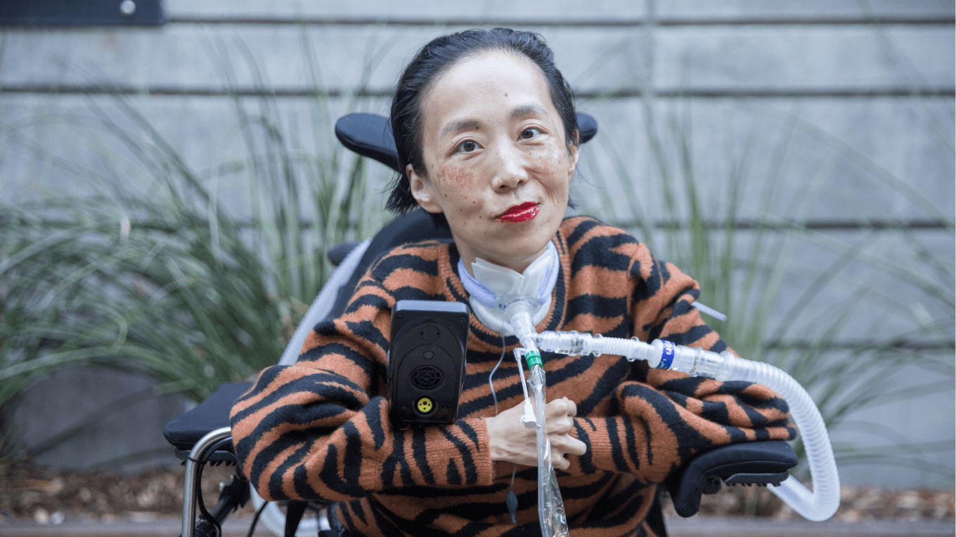 Photo of Alice Wong, an Asian American disabled woman in a power chair. She is wearing an orange and black tiger-striped sweater, black pants, a bold red lip color and a trach at her neck. In the background is a gray cement wall with greenery. Photo credit: Eddie Hernandez Photography.