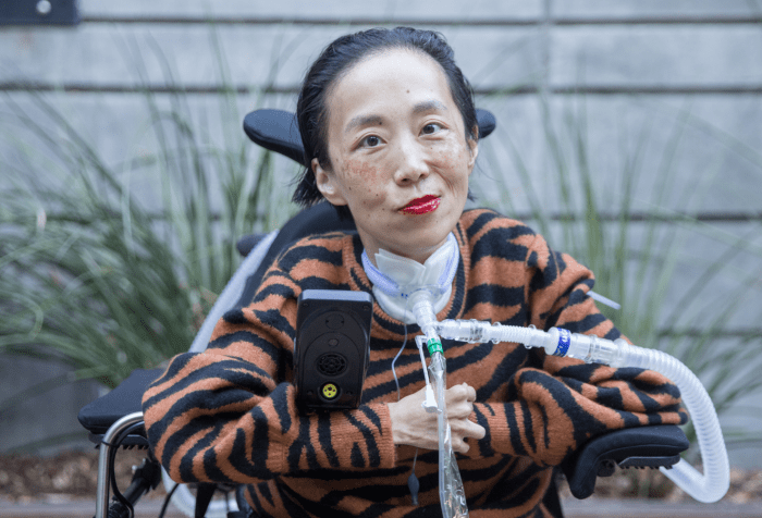 Photo of Alice Wong, an Asian American disabled woman in a power chair. She is wearing an orange and black tiger-striped sweater, black pants, a bold red lip color and a trach at her neck. In the background is a gray cement wall with greenery. Photo credit: Eddie Hernandez Photography.