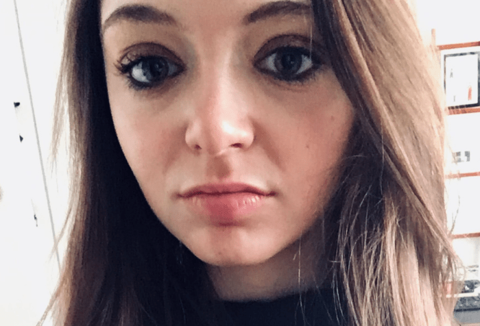 A woman with long wavy brown hair stands, smiling. Her face fills almost the entire photo frame. She has brown-colored eyes, brown eyeshadow, and pink lipstick while wearing a long sleeve black turtleneck shirt. Behind her is an office wall covered with small artworks and photographs.