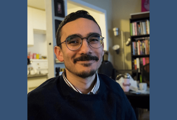 A dark blue background in the center, a close-up photo of Angelo, a man smiling with dark metal glasses and a short beard. He is wearing a navy blue sweater layered over a striped collared shirt.