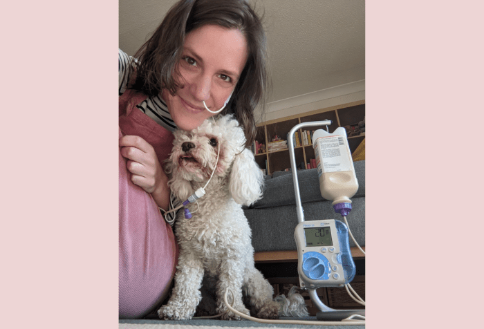 Background with a light pink background. Selfie of a white woman with dark hair and eyes with an NJ tube on her face, next to a white toy poodle, and with her feed hanging on a frame with a pump attached.