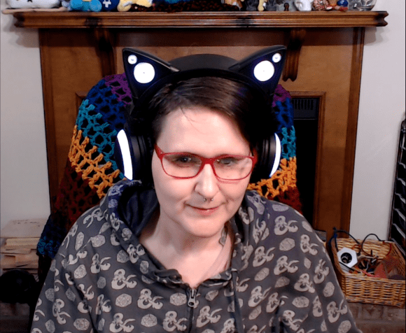 Young white woman wearing pink-framed eyeglasses and a hoodie. She is also wearing headphones that look like black cat ears. Behind her is a wooden mantle full of cute nerdy stuffed creatures.