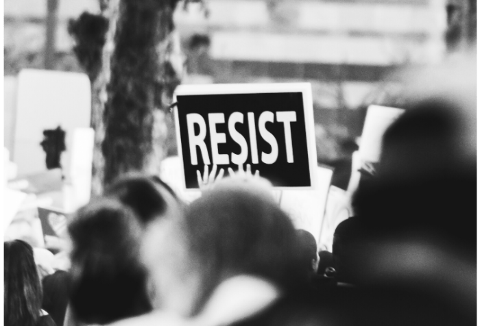 Black and white photo of an outdoor protest with one sign that says RESIST in white letters against a black piece of cardboard