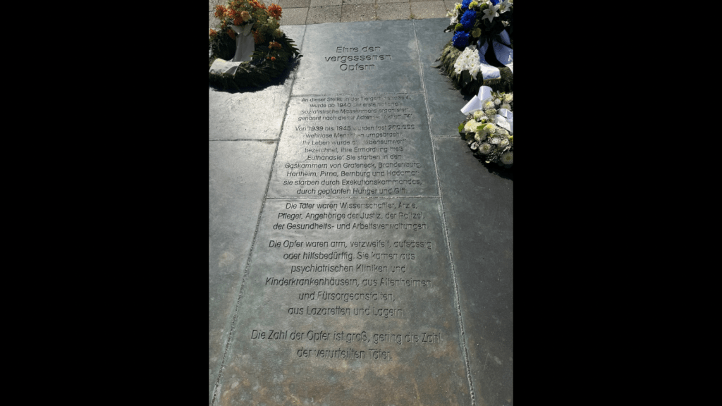 Graphic with a black background. Photo of a memorial plaque at Tiergartenstraße 4 in Berlin. The inscription reads: “Honor to the forgotten victims. At this site, at Tiergartenstraße 4, the first National Socialist mass murder was organized starting in 1940, named after this address: Aktion T4. From 1939 to 1945, almost 200,000 defenseless people were killed. Their lives were described as ‘unworthy of life,’ their murder called euthanasia. They died in the gas chambers of Grafeneck, Brandenburg, Hartheim, Pirna, Bernburg, and Hadamar; they died by firing squads, by deliberate starvation, and by poison. The perpetrators were scientists, doctors, nurses, members of the judiciary, the police, and of the health and labor administrations. The victims were poor, desperate, defiant, or in need of help. They came from psychiatric clinics and children’s hospitals, from nursing homes and welfare institutions, from military hospitals and camps. The number of victims was vast; the number of perpetrators convicted was very small.”