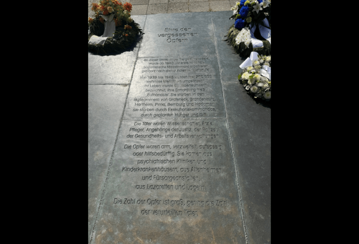 Graphic with a black background. Photo of a memorial plaque at Tiergartenstraße 4 in Berlin. The inscription reads: “Honor to the forgotten victims. At this site, at Tiergartenstraße 4, the first National Socialist mass murder was organized starting in 1940, named after this address: Aktion T4. From 1939 to 1945, almost 200,000 defenseless people were killed. Their lives were described as ‘unworthy of life,’ their murder called euthanasia. They died in the gas chambers of Grafeneck, Brandenburg, Hartheim, Pirna, Bernburg, and Hadamar; they died by firing squads, by deliberate starvation, and by poison. The perpetrators were scientists, doctors, nurses, members of the judiciary, the police, and of the health and labor administrations. The victims were poor, desperate, defiant, or in need of help. They came from psychiatric clinics and children’s hospitals, from nursing homes and welfare institutions, from military hospitals and camps. The number of victims was vast; the number of perpetrators convicted was very small.”