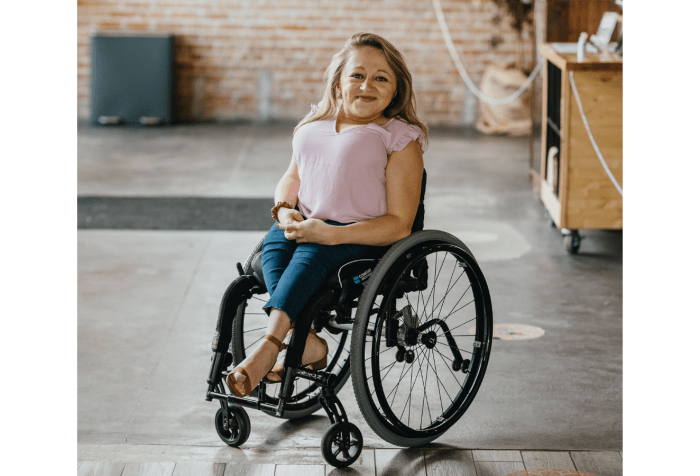 A white woman, Emily, with blonde, shoulder-length hair sits in a black wheelchair. She is short of stature. She is wearing a pink short-sleeved shirt and blue jeans with brown wedge heels. The ground is partially cement, partially wood. There is a brick wall behind her.