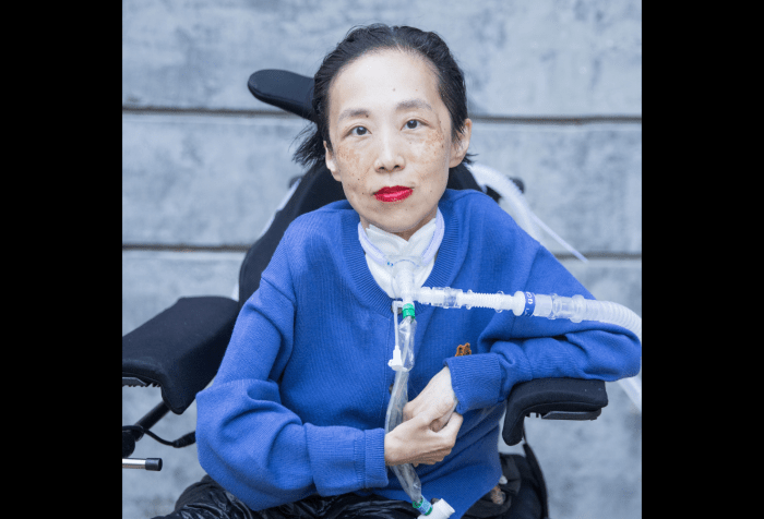Photo of Alice Wong in front of a cement wall. She is wearing a blue cardigan and sitting in a power chair. She is staring intently at the camera, wearing a bold red lip color, and a trach at her neck. Photo credit: Eddie Hernandez Photography.