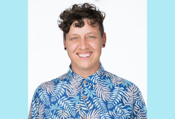 Graphic with a light blue background with a photo of a smiling non-binary white person with short curly brown hair wears black earring plugs and a bright blue button up shirt with colorful fern fronds printed on it.