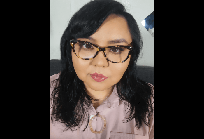 Picture of Johnnie Jae, black hair, bangs brushed to the right side of her face. She is wearing glasses that are slightly too big for her head, nude pink lipstick, a dusky pink shirt with a necklace featuring a round citrine pendant.