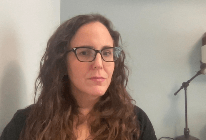 headshot of Justine Barron, a white woman with long wavy brown hair and black glasses, seated in front of a desk lamp.