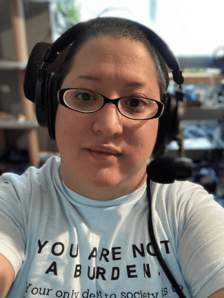 Alejandra is a light-skinned Latina woman with close cropped dark hair. In a close-up selfie, she wears a headset with a microphone, glasses with narrow black frames, and a short-sleeved, light blue shirt where the first part of a quote by Lydia X. Z. Brown is visible: YOU ARE NOT A BURDEN.