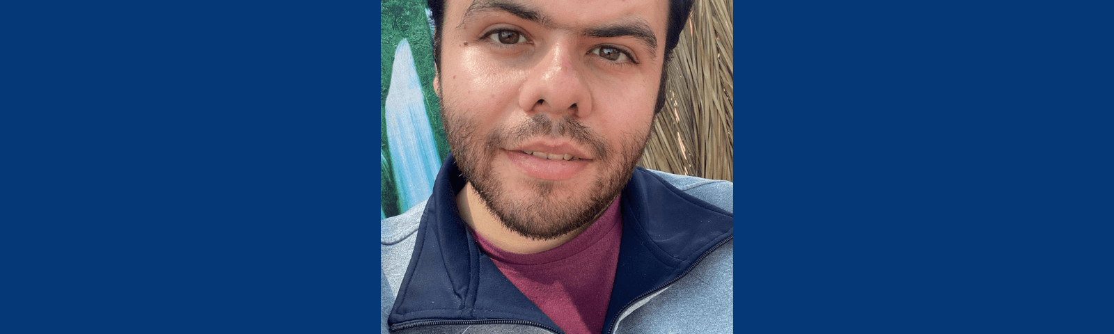 Graphic with a dark blue background. In the center is a close-up of Khaled outside in the sun, dark brown hair combed up, slight brown beard and hazel-brown eyes staring into the camera. He wears a slight smile and a blue and gray zip up with maroon shirt underneath. 