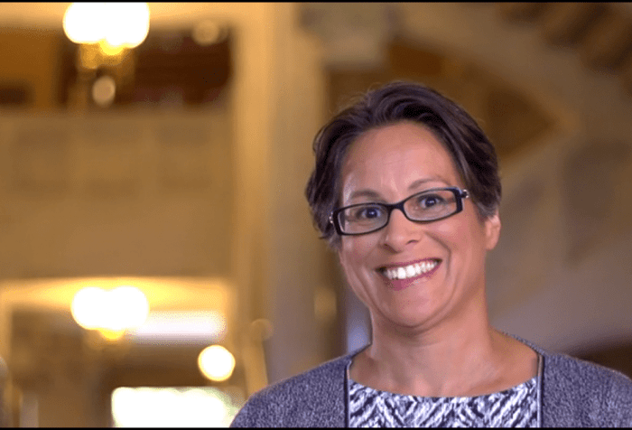 A head and shoulders picture of a woman with short brown/gray hair, glasses, and a big smile standing in front of a column in a government building. The picture is off center and there are blurred lights in the background. She is wearing a gray patterned sweater over a gray, blue and black patterned dress.