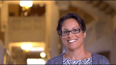 A head and shoulders picture of a woman with short brown/gray hair, glasses, and a big smile standing in front of a column in a government building. The picture is off center and there are blurred lights in the background. She is wearing a gray patterned sweater over a gray, blue and black patterned dress.