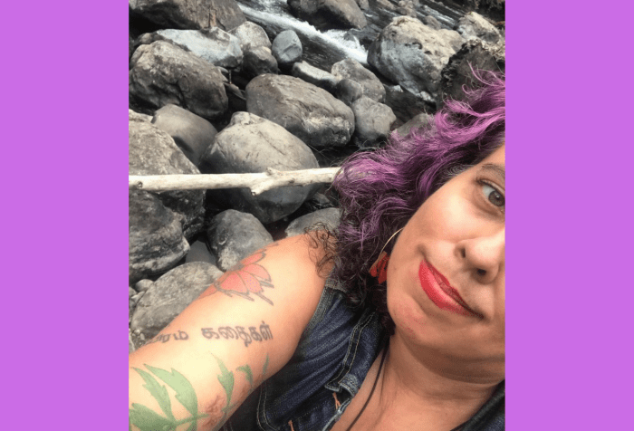 Graphic with a light purple background with a photo of a middle aged mixed race Sri Lankan, Irish and Romani femme, diagonially, in front of a wild river with lots of rocks. Leah has violet, brown and silver curly hair, peachy tan sand colored skin and red lipstick (Stila Stay All Day lipstick in Beso, aka "the lipstick AOC wears.") Their clothes are mostly not visible except for a blue denim vest. You can see their sleeve of tattoos, including cosmos flowers, a motherwort plant and letters in Tamil. They are smiling with bashful pride and satisfaction."