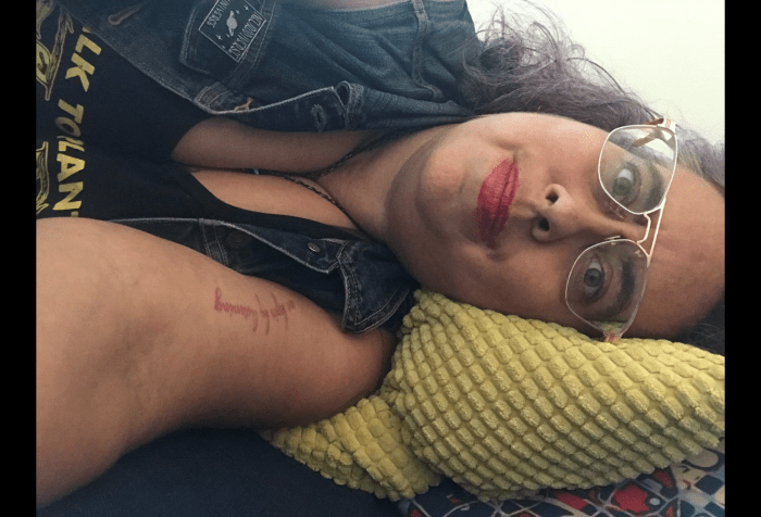 Graphic with a black background with a photo of a 40ish mixed race Sri Lankan, Irish and Galician nonbinary  femme with curly brown silver and purple hair, lying on a couch looking at the viewer horizontally. They have rose gold aviator frames, thick eyebrows, red lipstick and sand colored skin, and are looking at the viewer with a kind of tired but hopefully crip wonder. They wear a blue denim vest with a pin that says Neurodivergent Universe above a pink and blue image of a ringed planet, and a black tank top with yellow lettering that read Talk To Plants, Not Cops is barely visible. They have a tattoo of the words "We begin by listening" in magenta cursive script on their left arm.