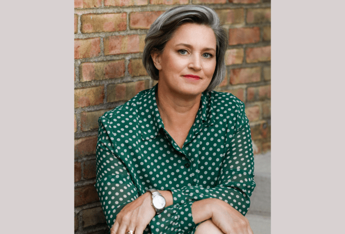 Graphic with a taupe background. In the center is a photo of Margaret Price, a white genderqueer femme, wears a green polka-dot dress and sits in front of a brick wall. Her chin-length hair is gray and her face is smiling slightly.