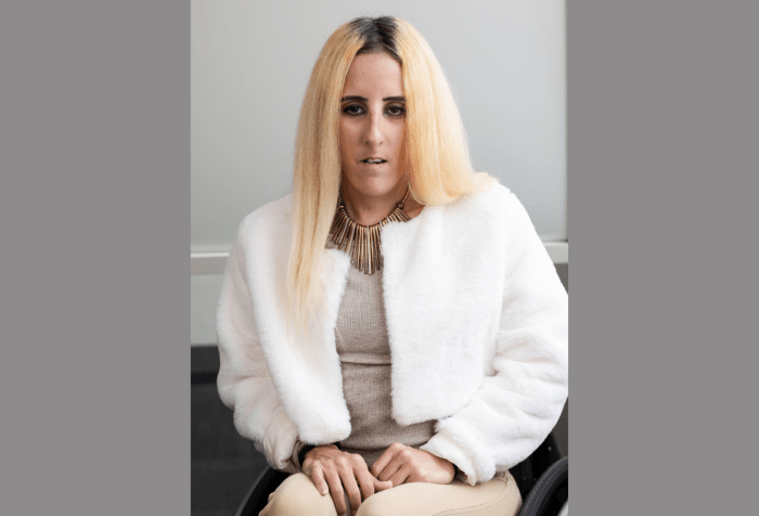 A headshot of a woman with brown color eyes and olive-tan skin complexion with long, straight blonde hair, wearing a baige top with a white jacket and a gold necklace. 