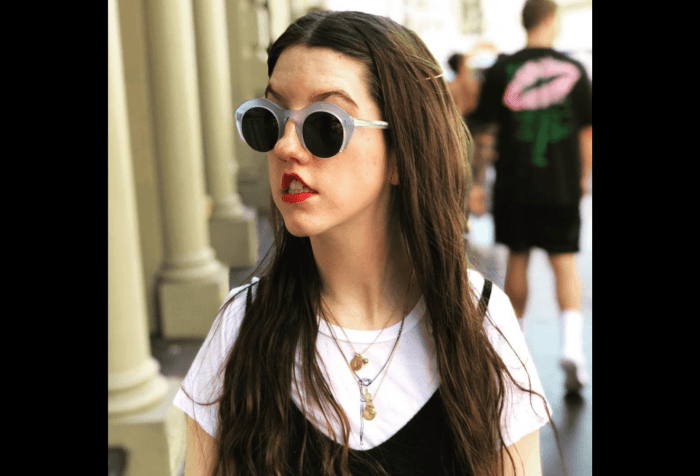 A young, white disabled woman with long dark hair, standing outside and looking off to the side, wearing light blue sunglasses and a white t-shirt with a small black walking stick design and a black sleeveless jumpsuit over the shirt.