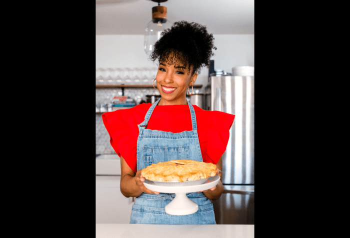 Graphic with a black background and a photo of Maya-Camille Broussard, a young Black woman with curly black hair who is wearing gold hoop earrings. She is wearing a bright red top with ruffled short sleeves and a denim apron. She is holding a white ceramic cake stand with a pie that has a golden crust.