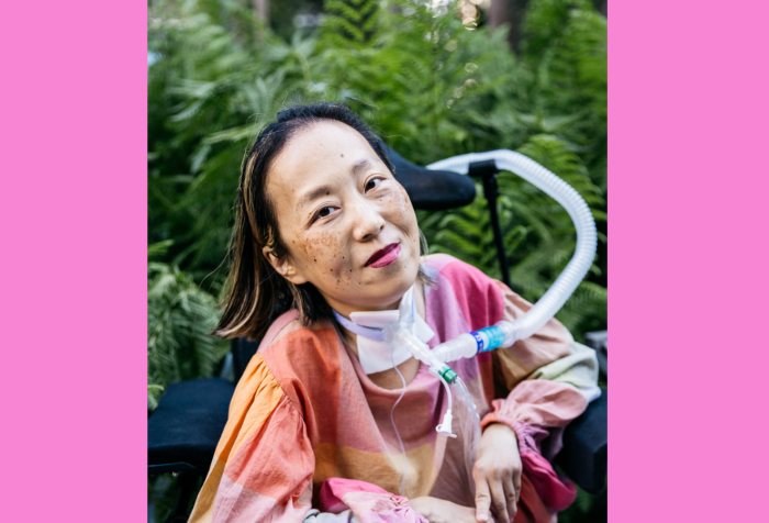 Graphic with a pink background. In the center is a photo of Alice Wong, Asian American woman in a wheelchair with a tracheostomy at her neck connected to a ventilator. She’s wearing a pink plaid shirt, pink pants, and a magenta lip color. She is smiling and behind her are a bunch of large fern-like plants. Photo credit: Allison Busch Photography.