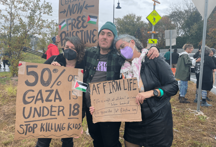 Image of Leah and two friends at a protest. One friend is a white, Greek/Irish femme person in her 40s with greying undercut brown hair wearing a black KN95 mask, they are holding a sign that says 50% OF GAZA IS UNDER 18 STOP KILLING KIDS. The second person is a Syrian masculine person wearing a green hat, a green and black flannel over a black t shirt with lilac lettering, holding a sign that reads CEASEFIRE NOW: FREE PALESTINE with two red black green and white Palestinian flags. Leah stands to the right leaning on their friend with their head on their shoulder, wearing a black leather jacket, black leather skirt, pink and white keffiyeh and lilac KN95 mask, holding a sign that reads, "Affirm life." Protestors can be seen in the background at a rotary in mid fall by a tree
