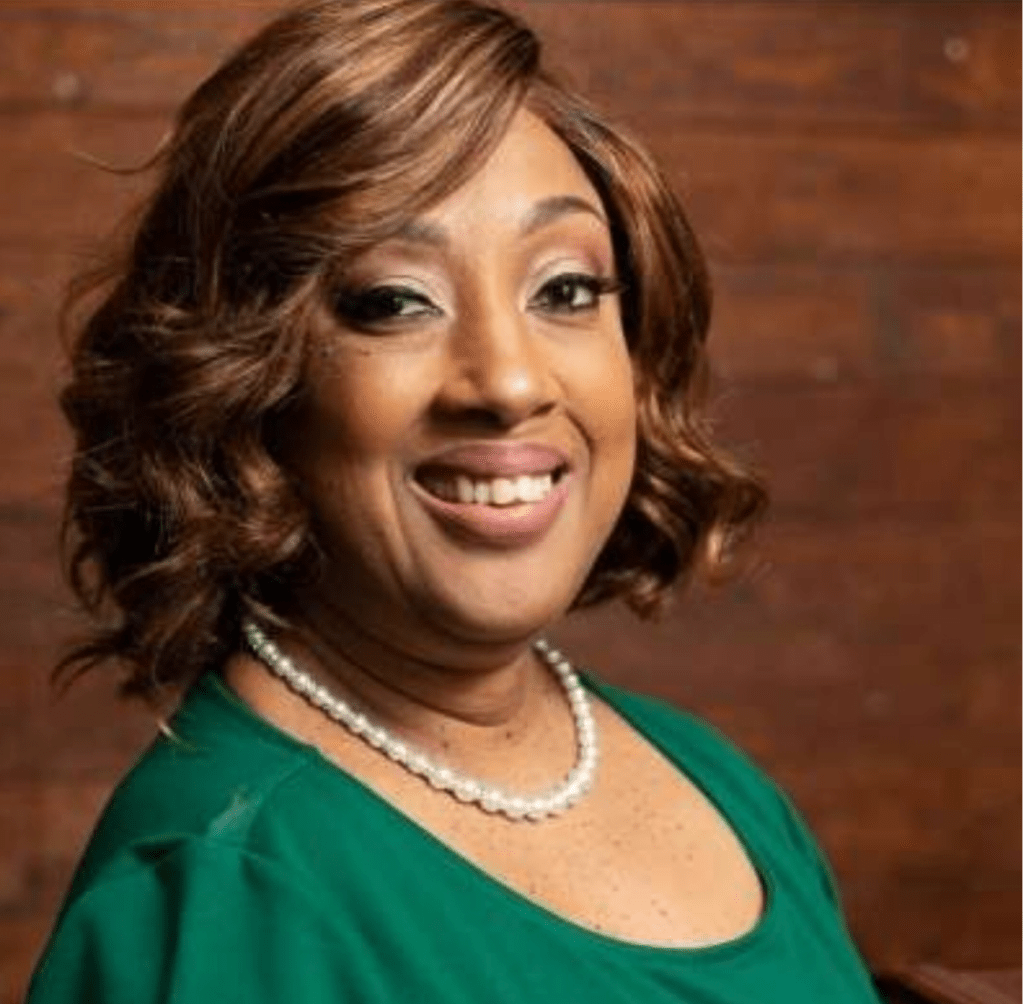 A photo of Rickii, Black woman with curled medium-length brown hair smiling at the camera. She is wearing pearl necklace and green shirt.