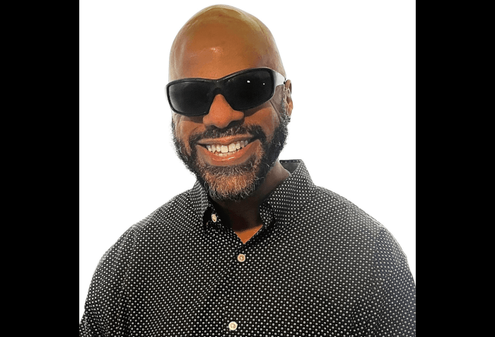 Graphic with a black background featuring a photo of Thomas Reid, a brown skin Black man with a clean-shaven bald head and full neat beard,  smiles into the camera. He wears dark shades and a black and white checkered button up shirt.
