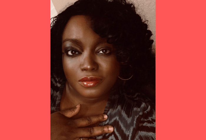 Graphic with a coral background and a head and shoulders picture of Tinu Abayomi-Paul, against a light brown walk. She is a dark skinned woman with chin length curly black hair. She has on red lipstick and her left gold colored hoop earring is visible. She is not smiling, but looks content. She has a hand lightly pressed against her chest and is wearing a dark blue, light blue and black blouse patterned in zigzag stripes. She's looking directly into the camera.