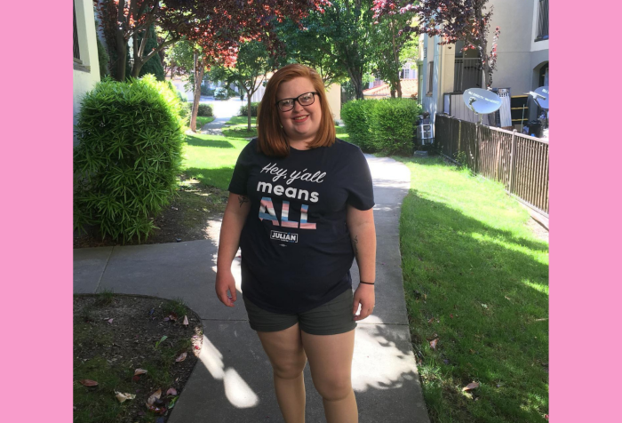 Graphic with a pink background and a A standing picture of Tory Cross, a white woman with red hair and black glasses, smiling and wearing green shorts a blue shirt that says “Hey Ya’ll Means All” with hey y’all means in white font and All in stripes of blue, pink and white. There is a sidewalk and greenery behind her.