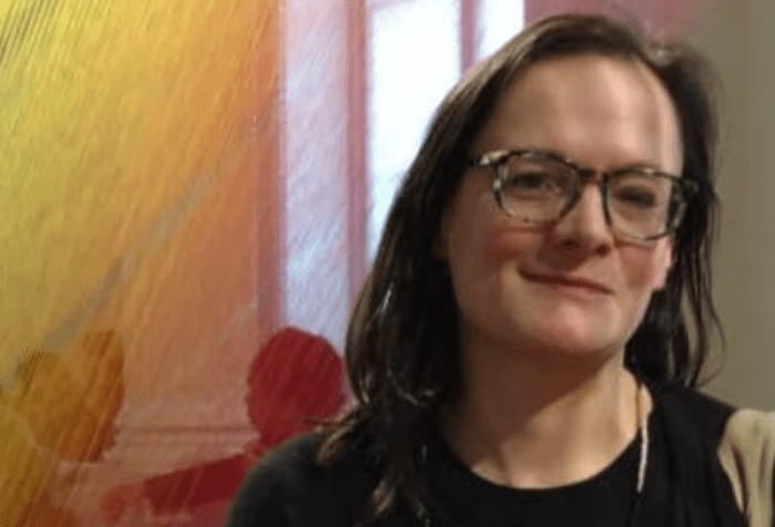 Cropped photo of Bess Williamson, a white woman with brown hair and glasses, standing in front of a window and a rainbow thread artwork by Gabriel Dawe.