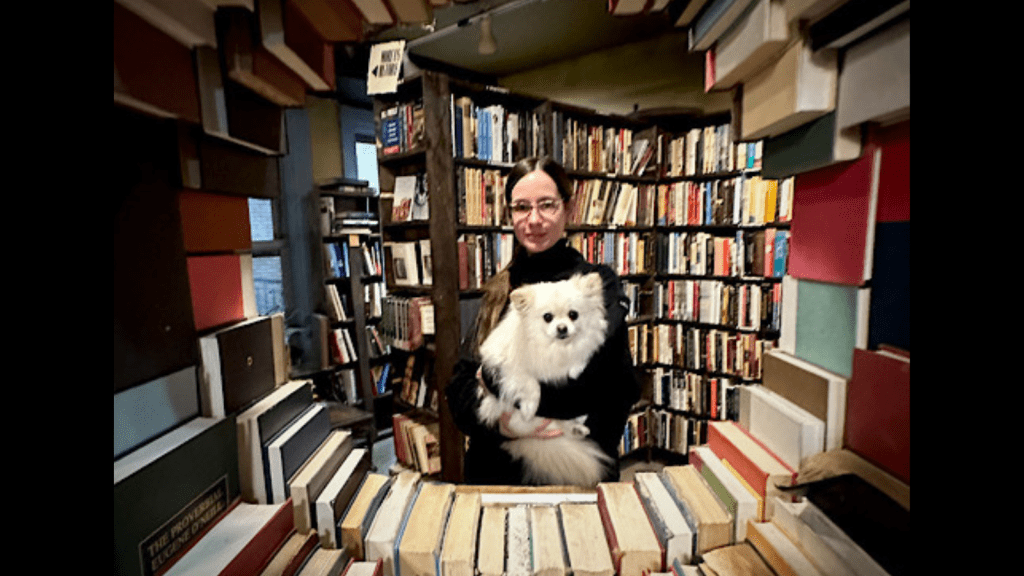 A white woman wearing glasses and a black sweater smiles while holding a small, fluffy white dog in her arms. They are surrounded by tall shelves filled with books, and are gazing together at the camera through a circular “window” opening in a sculptural wall made of hardcover books in an array of colors.